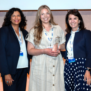 Photo of three women. The woman in the middle is holding a trophy. They are all smiling.