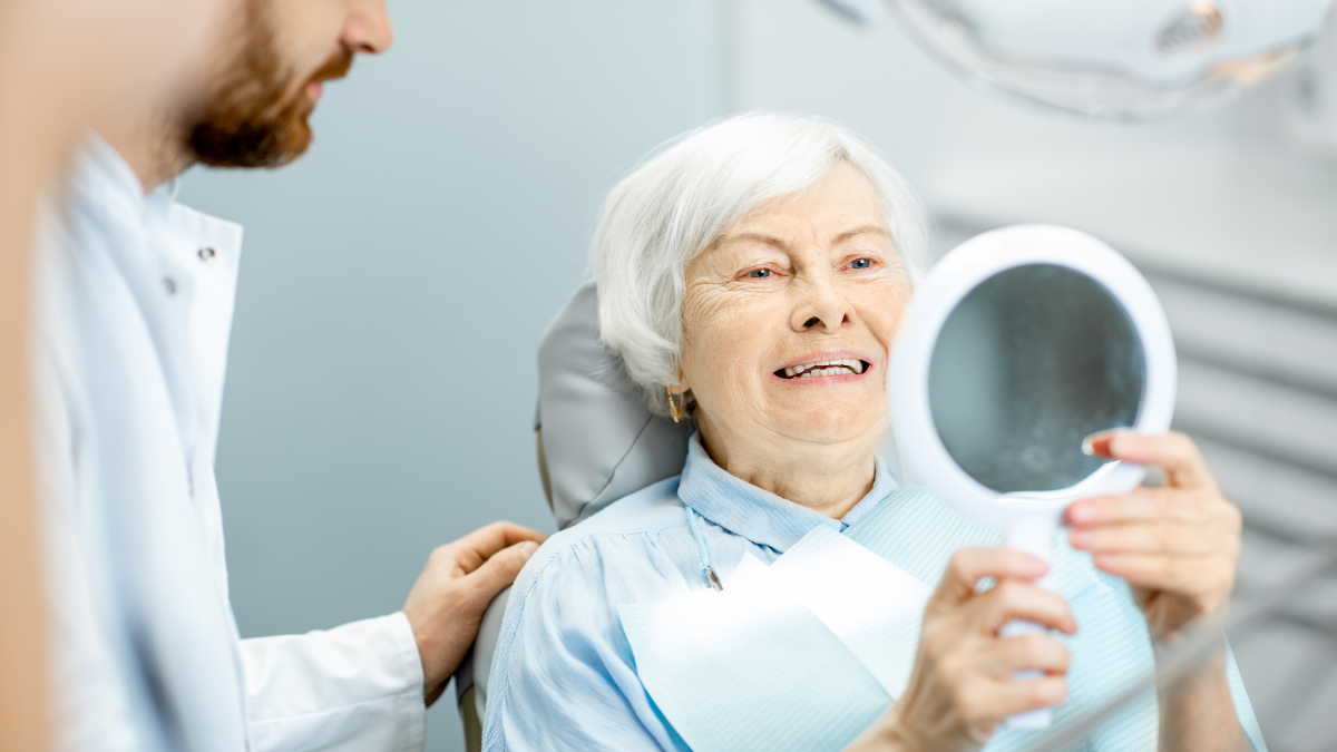 Elderly patient sat in dentist chair holding mirror and looking at teeth