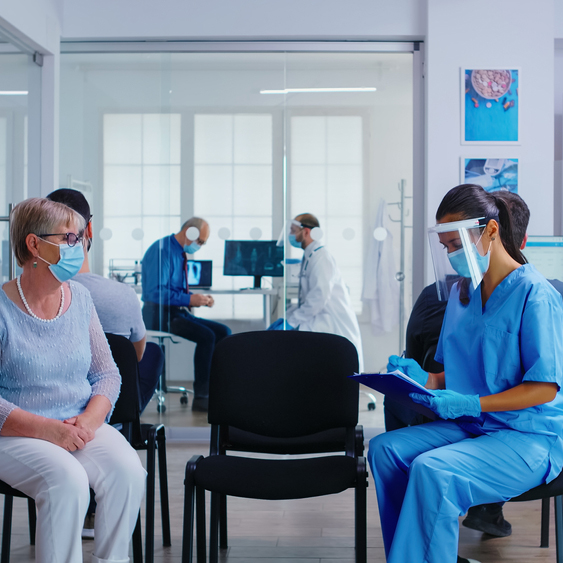 Nurse and senior patient going in hospital examination room from waiting area. Doctor inviting old woman for consultation in office. Wearing mask against coronavirus.