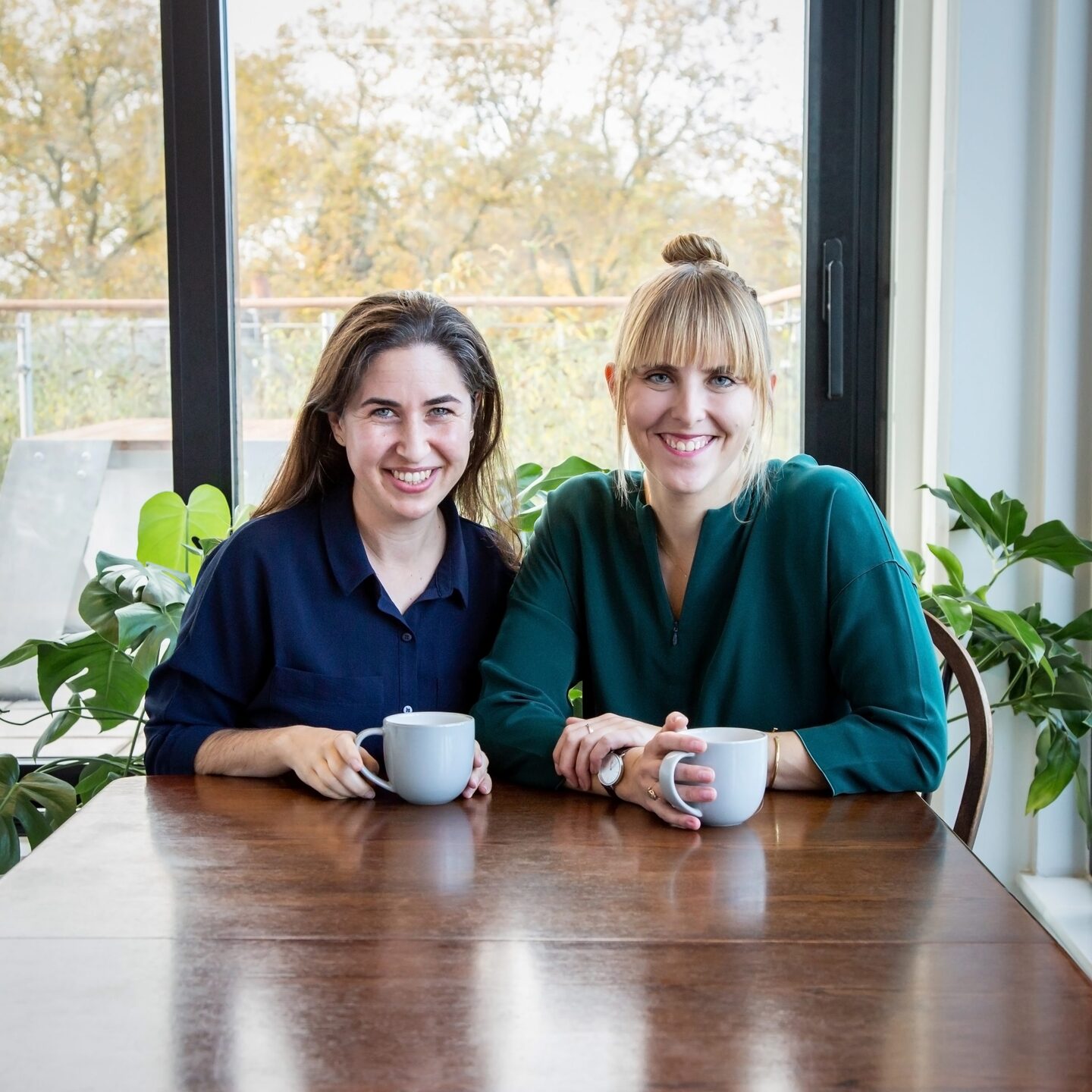 Two women sat at table with hot drinks smiling at camera