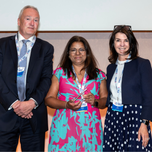Photo of a lady wearing a blue and pink dress holding a trophy. She is stood in the middle of a man wearing a dark suit and a woman wearing a dark blue jacket and blue skirt with white polka dots. They are smiling.