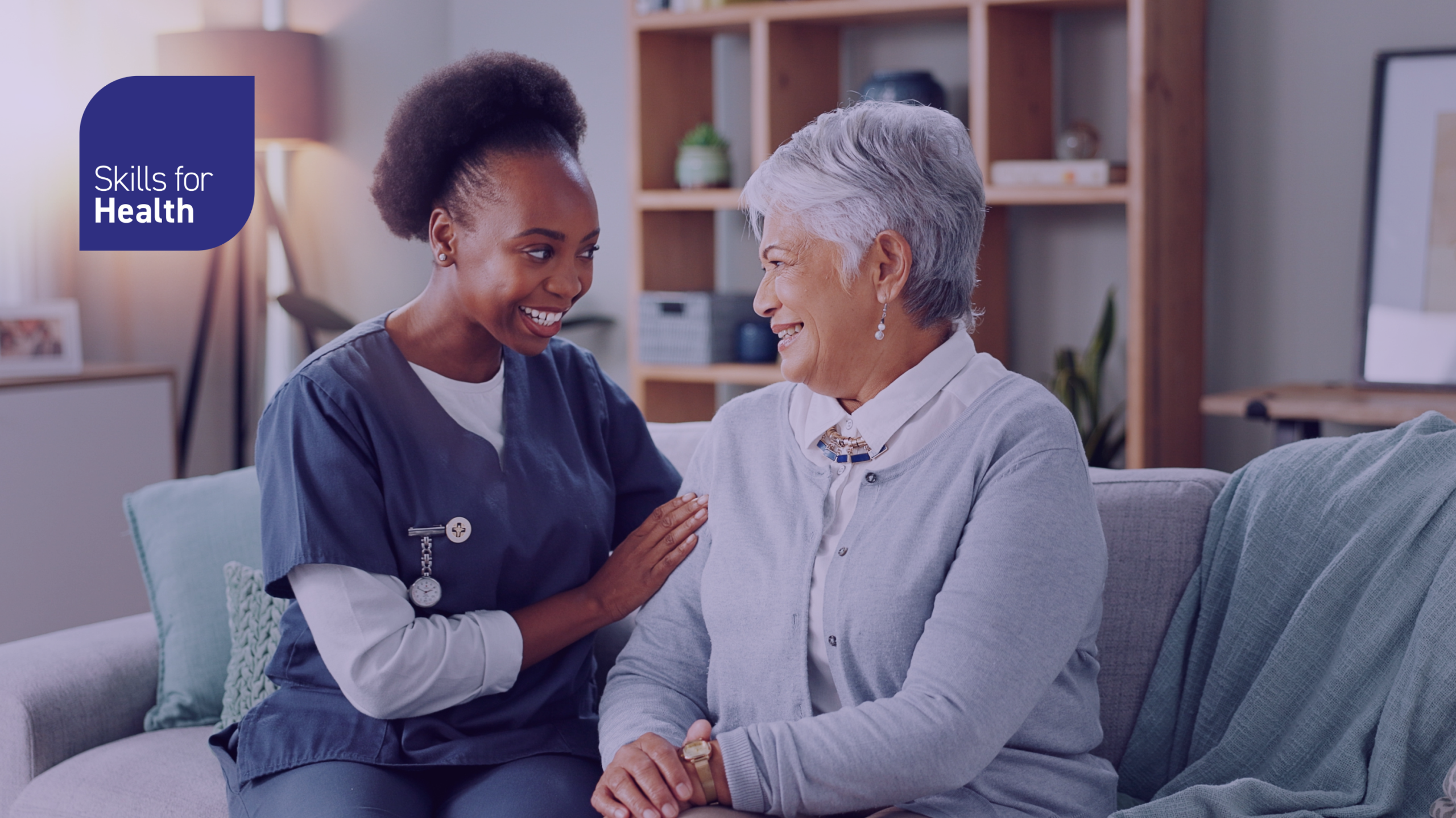 Medical health worker in scrubs sat on a sofa with an elderly lady they are smiling