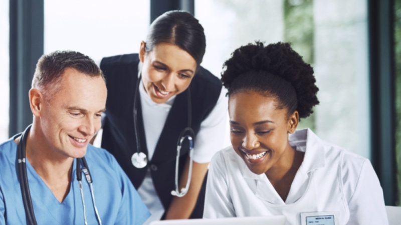 Healthcare team in a bright office around a laptop, with doctors and nurses smiling and listening as one clinician points at the screen.