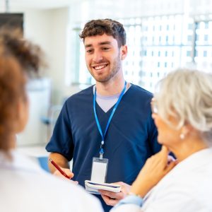 Young male nurse smiling and taking notes while talking with medical team in a hospital hallway