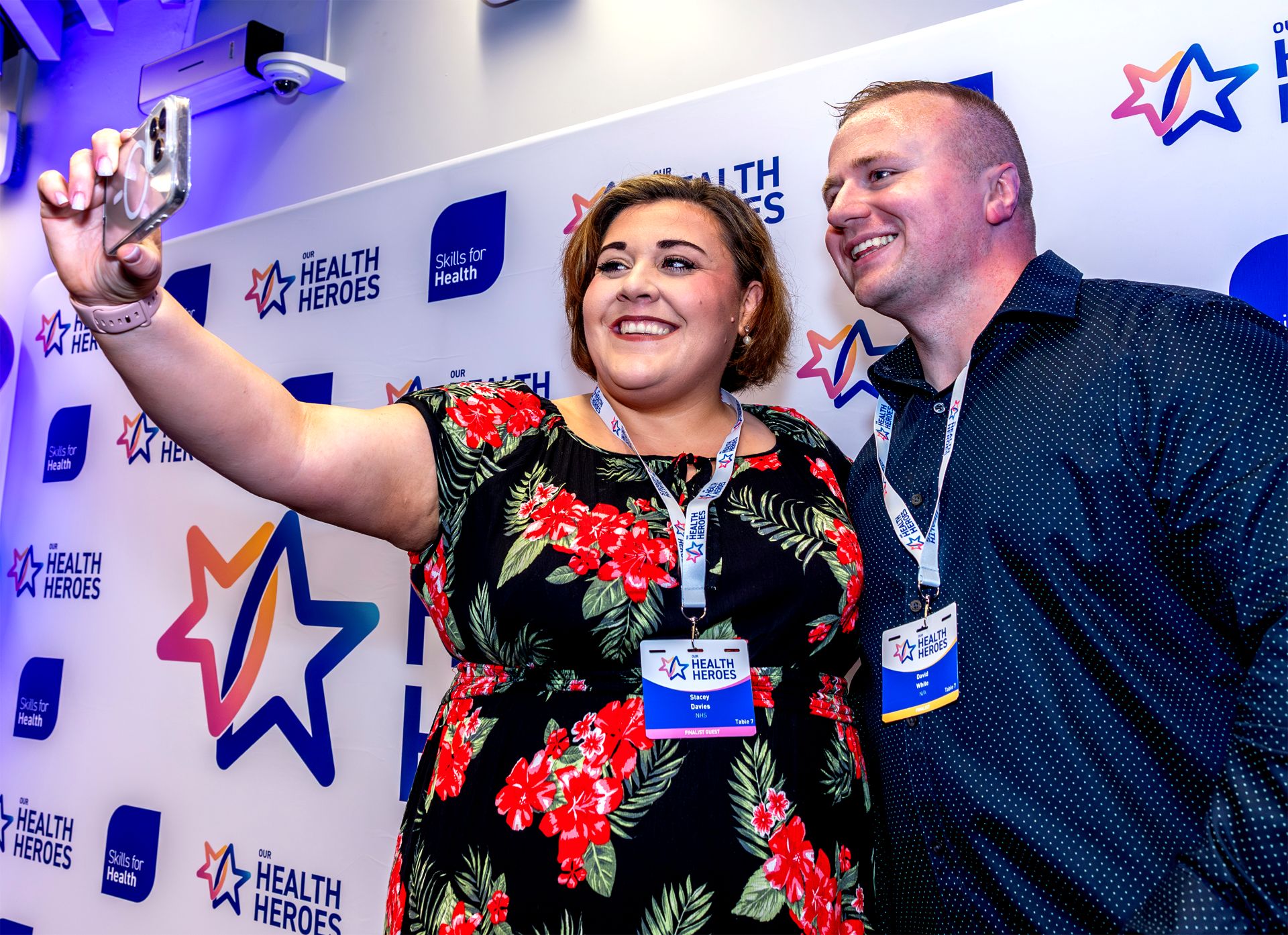 Smiling man and woman taking a selfie in front of a branded back drop.